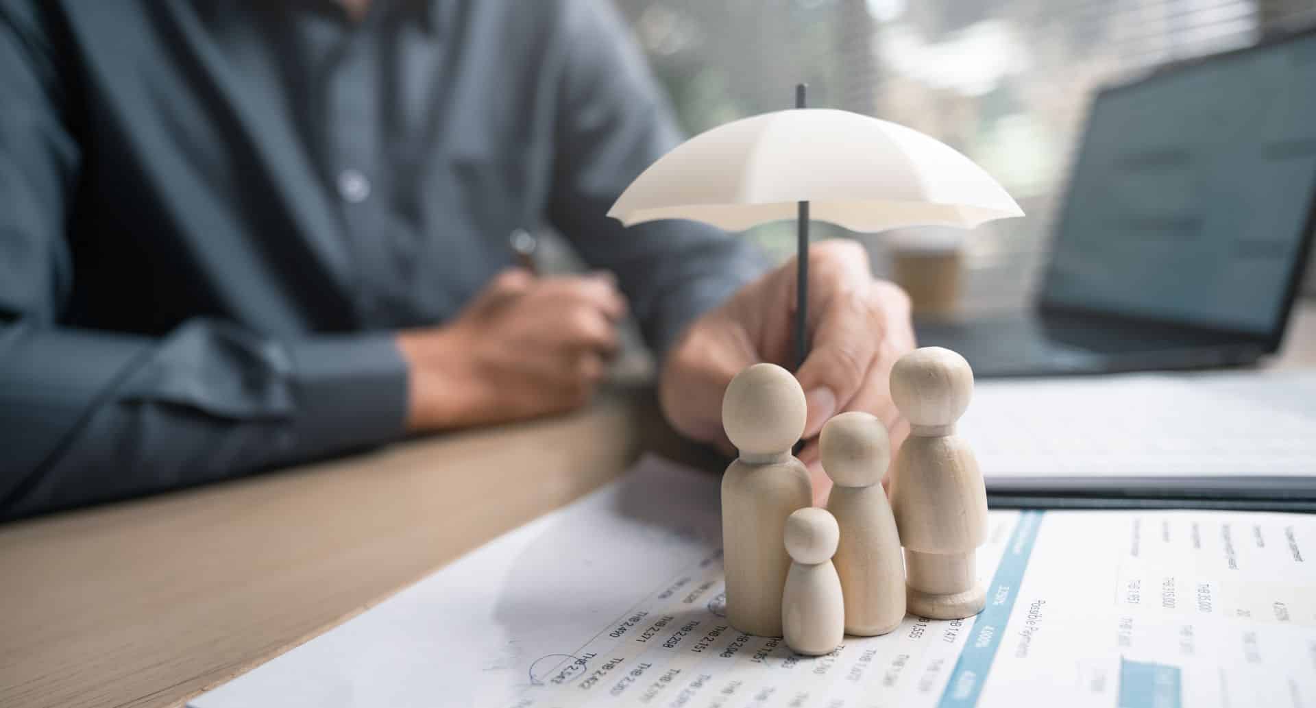 Family insurance plan protect. Help medicine protection. Father mother children insurance. A man is holding a white umbrella over a family of three wooden figures